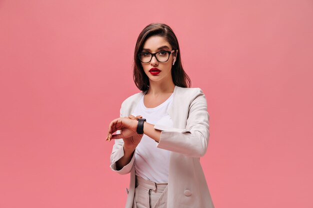 La signora di affari in occhiali guarda l'orologio su sfondo rosa. Bella ragazza seria con labbra rosse in abito elegante beige in posa.