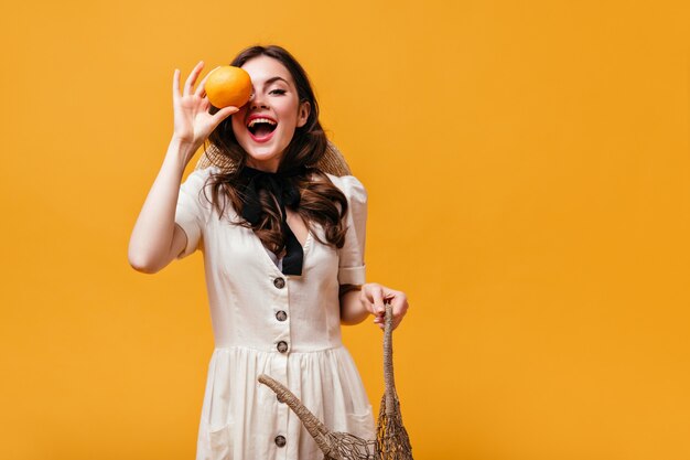 La signora in abito bianco ride, si copre gli occhi con l'arancio e tiene la borsa ecologica su sfondo arancione.