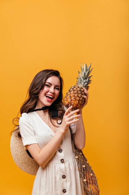 La signora in vestito di cotone sta tenendo l'ananas. Donna con capelli ondulati in posa con la borsa della spesa su sfondo arancione.