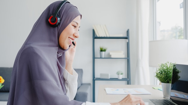 La signora musulmana asiatica indossa le cuffie guarda il webinar ascolta il corso online comunica tramite videochiamata in conferenza a casa.