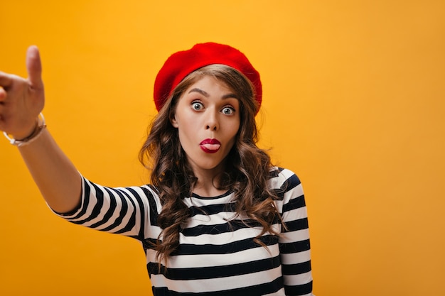 La signora positiva in berretto rosso mostra la lingua e fa selfie. Donna divertente in camicetta alla moda a righe e berretto rosso in posa su sfondo arancione.