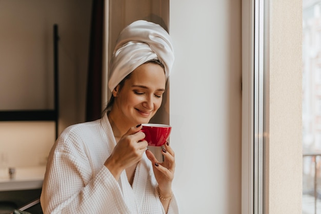 La signora sorridente nella sua veste mattutina inala con piacere l'aroma del caffè Donna in asciugamano bianco sulla testa che tiene la tazza rossa