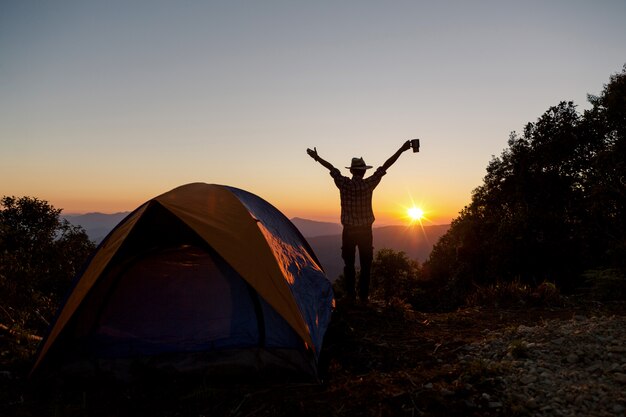 La siluetta dell&#39;uomo felice con la tazza di caffè della tenuta resta vicino alla tenda intorno alle montagne