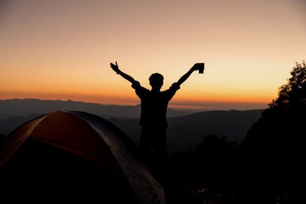 La siluetta dell&#39;uomo felice con la tazza di caffè della tenuta resta vicino alla tenda intorno alle montagne