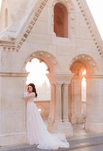La sposa attraente in tenero vestito alla moda è in piedi vicino alla colonna di pietra in una calda giornata estiva