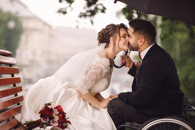 La sposa e lo sposo sulla sedia a rotelle si siedono baciare sulla panchina nel parco