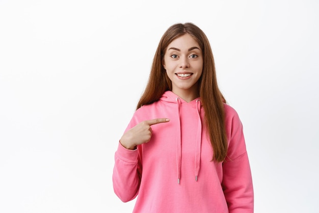 La studentessa adolescente carina indica se stessa e sorride volontaria per partecipare sceglimi gesto autopromozione abilità personali si erge su sfondo bianco