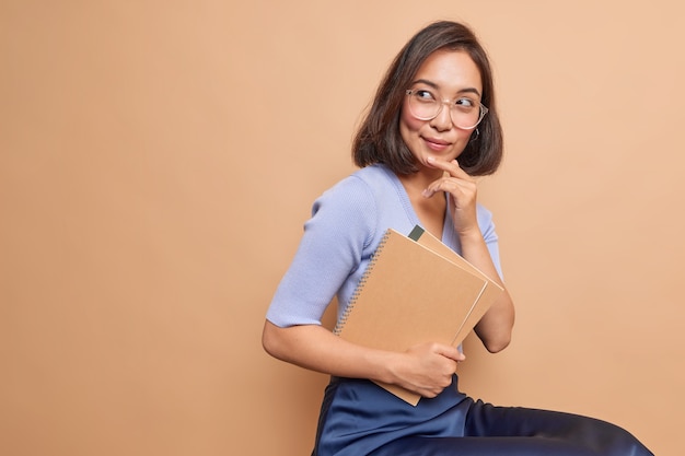 La studentessa asiatica premurosa porta i quaderni a spirale torna a scuola pensa come migliorare la sua conoscenza indossa occhiali abiti casual si siede spazio copia vuoto al coperto sulla parete beige