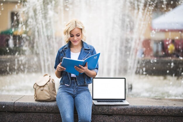 La studentessa bella ragazza bionda lavora sul suo computer portatile e legge il libro vicino alla fontana della città nel corso della giornata