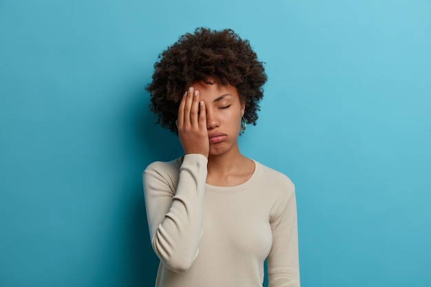 La studentessa esausta oberata di lavoro fa il palmo della mano, chiude gli occhi per la stanchezza, trascorre la notte insonne a prepararsi per gli esami, posa infelice al coperto, indossa un maglione bianco casual, isolato sul muro blu