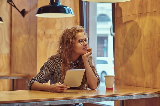 La studentessa riccia rossa intelligente e pensierosa tiene una tavoletta digitale mentre è seduta a un tavolo nella mensa del college durante una pausa.