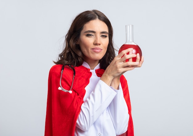 La superdonna soddisfatta in uniforme del medico con il mantello rosso e lo stetoscopio tiene il liquido chimico rosso nel pallone di vetro isolato sulla parete bianca