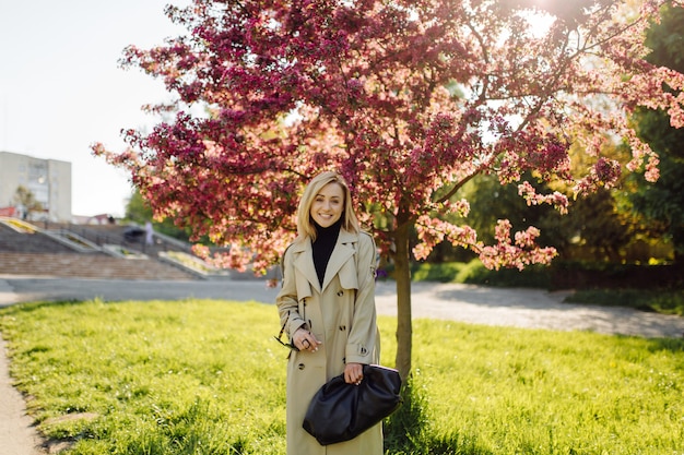 La trincea wearind della donna bionda caucasica sorride felicemente sulla soleggiata giornata di primavera fuori a piedi nel parco