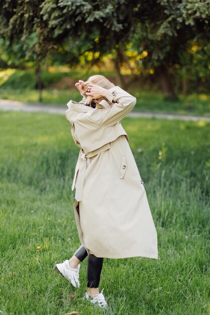 La trincea wearind della donna bionda caucasica sorride felicemente sulla soleggiata giornata di primavera fuori a piedi nel parco