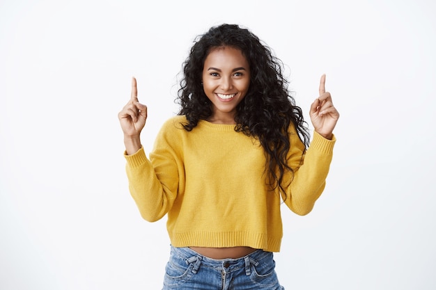 La tua pubblicità quassù. Allegra attraente incoraggiata giovane donna afro-americana con acconciatura riccia, indossa un maglione giallo sorridente fiducioso, rivolto verso l'alto, promuove lo spazio della copia superiore