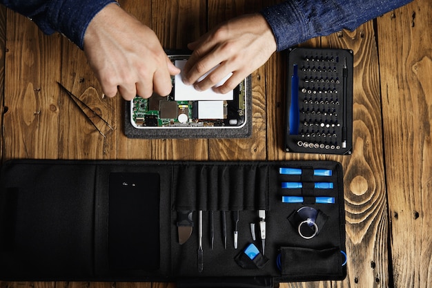 La vista dall'alto delle mani funziona sul gadget elettronico rotto per ripararlo vicino alla borsa degli attrezzi e sul tavolo di legno nel negozio di servizi