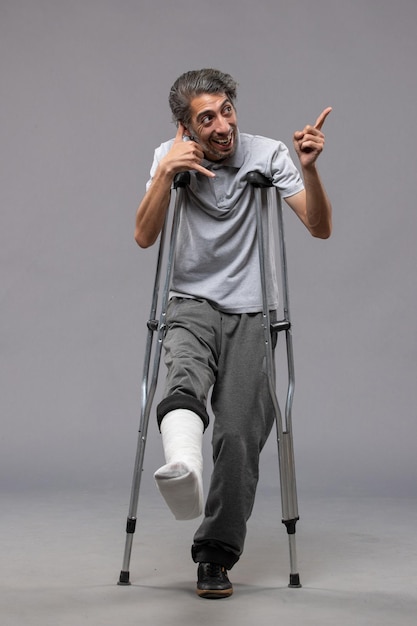 La vista frontale del giovane maschio con le stampelle a causa del piede rotto sul muro grigio disabilita la gamba rotta dell'incidente