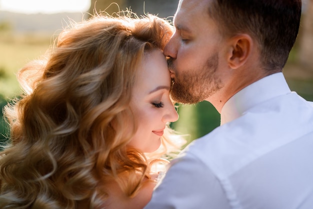 La vista frontale dell'uomo barbuto sta baciando la ragazza bionda con la pettinatura e il trucco sulla fronte all'aperto il giorno soleggiato