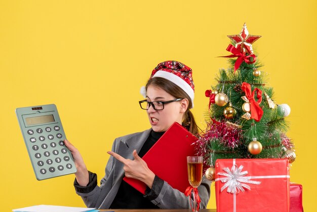 La vista frontale ha vagato la ragazza con il cappello di natale che si siede al tavolo guardando l'albero di Natale del calcolatore e il cocktail dei regali