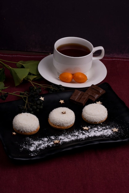 La vista laterale dei biscotti con i pezzi del cioccolato dei fiocchi della noce di cocco su un bordo nero è servito con tè su oscurità
