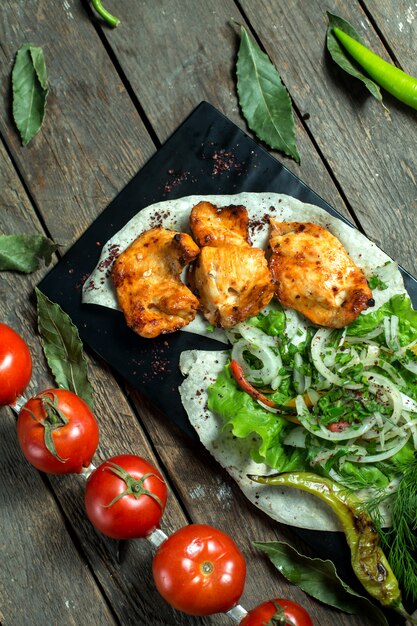 La vista laterale del kebab di pollo è servito con il pomodoro ed il pepe grigliati erbe fresche delle cipolle sul bordo nero