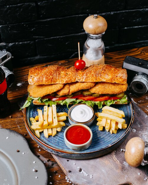 La vista laterale del panino con l'insalata delle pepite di pollo lascia i sottaceti e la salsa serviti con le patate fritte sulla tavola di legno