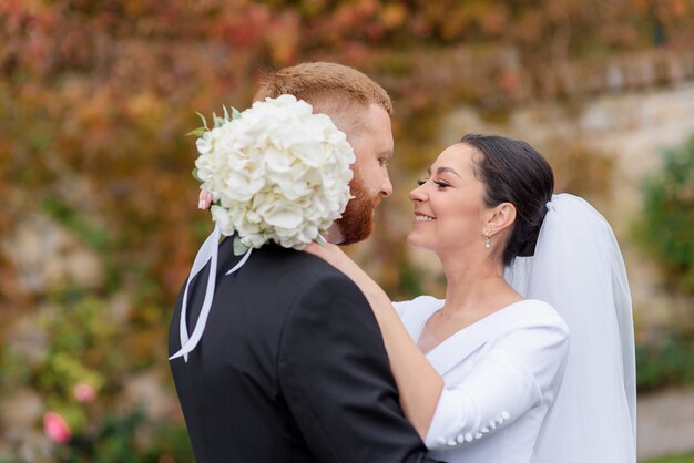 La vista laterale della bella sposa bruna sorride e abbraccia il suo sposo dai capelli rossi