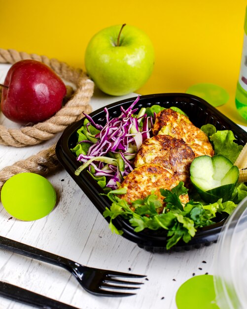 La vista laterale della cotoletta della patata e dei broccoli è servito con l'insalata di cavolo in una scatola della consegna sulla tavola di legno