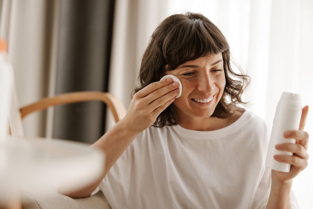 La vista laterale della donna sorridente con lozione per il viso chiude gli occhi