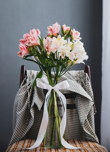 La vista laterale di un mazzo di alstroemeria rosa e bianco di colore fiorisce in un vaso di vetro su una sedia di legno al fondo grigio della parete