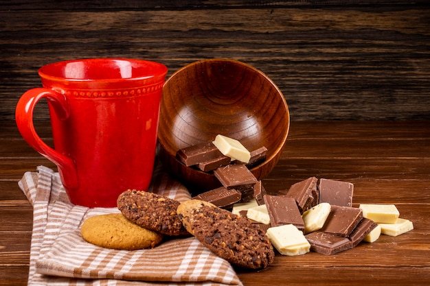 La vista laterale di una tazza con i biscotti di farina d'avena del tè e la cioccolata scura e bianca collega i pezzi sparsi dalla ciotola di legno su fondo rustico