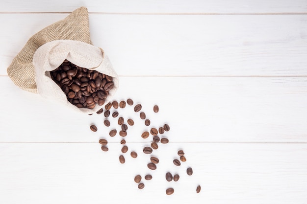 La vista superiore dei chicchi di caffè ha sparso da un sacco su fondo di legno bianco con lo spazio della copia