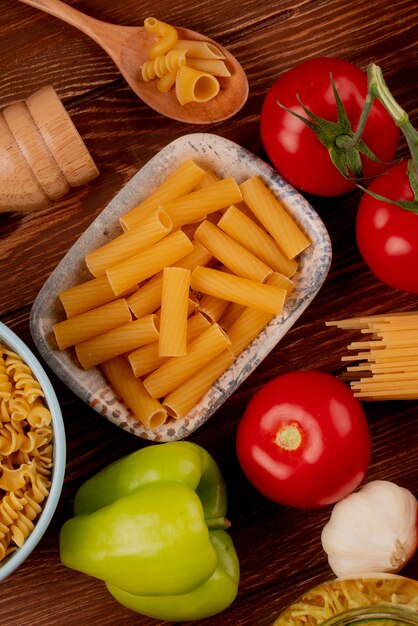 La vista superiore della pasta di ziti in ciotola con spaghetti e il rotini scrive in pepe di aglio del pomodoro del sale e del cucchiaio della ciotola su superficie di legno