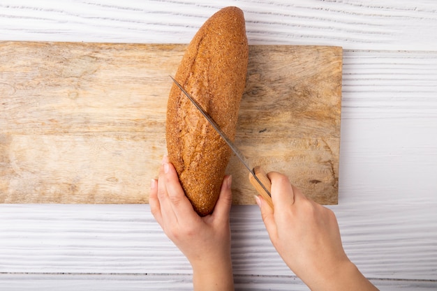 La vista superiore delle mani della donna che tagliano le baguette con il coltello sul tagliere su fondo di legno