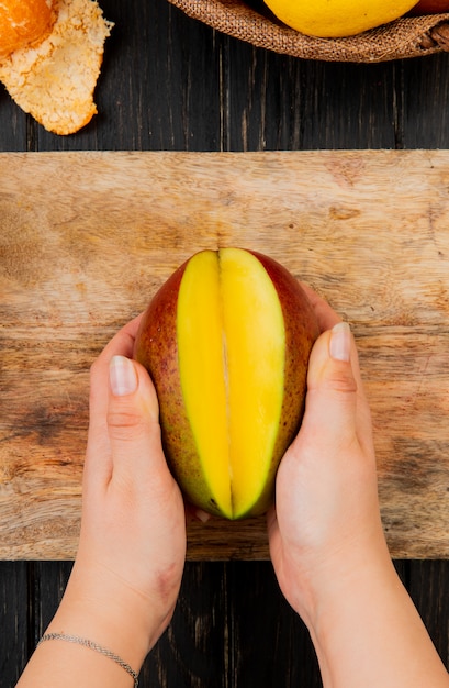 La vista superiore delle mani della donna che tengono la metà ha tagliato il mango sul tagliere sulla tavola di legno