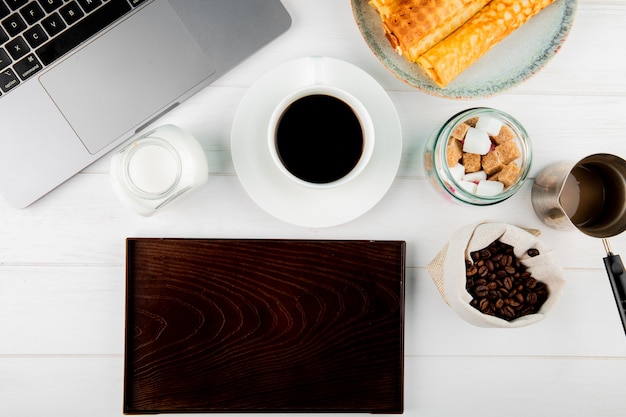 La vista superiore di una tazza di caffè con il wafer rotola i chicchi di caffè in un computer portatile del sacco e un bordo di legno su fondo bianco