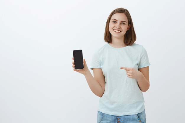 La vita è cambiata dopo questo telefono. Ritratto di donna affascinante felice dall'aspetto amichevole con capelli castani corti in maglietta leggera casual che mostra lo schermo del cellulare e che punta allo smartphone sorridente