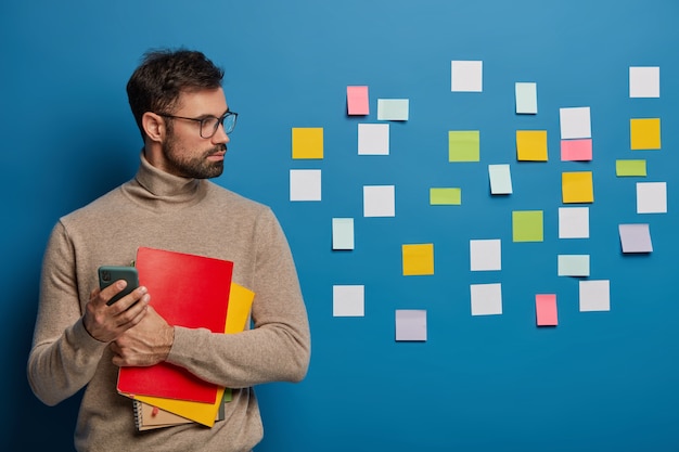 La vita in su di un uomo barbuto ha un pensiero creativo, blocchi appunti colorati bloccati sul muro blu, tiene il blocco note a spirale