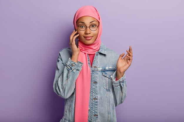 La vita in su di una donna musulmana gode di conversazioni con lo smartphone, essendo un utente avanzato del dispositivo moderno, indossa hijab rosa e giacca di jeans, utilizza la connessione Internet pubblica, isolata sul muro viola