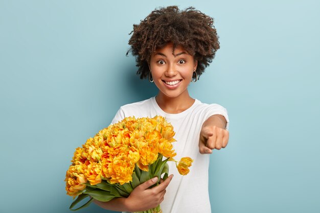 La vita sul colpo di felice bella ragazza afro punta direttamente alla telecamera con il dito anteriore, indossa una maglietta bianca casual