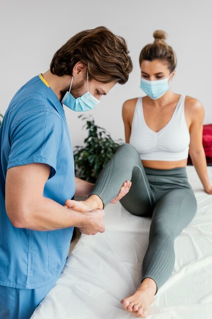 Lato del terapista osteopatico maschio con mascherina medica che controlla la caviglia del paziente femminile