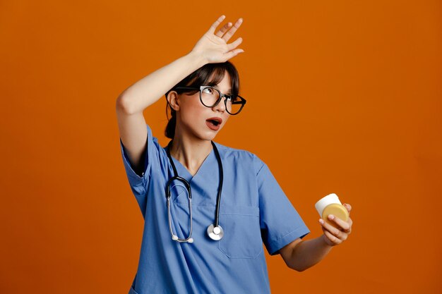 Latta stanca della holding della giovane dottoressa della pillola che indossa uno stetoscopio fitth uniforme isolato su sfondo arancione