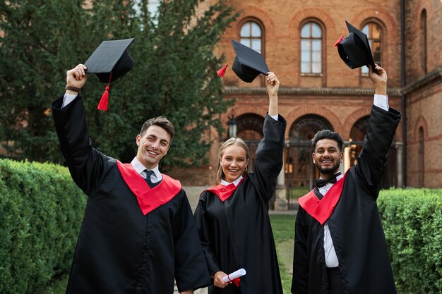 Laureati multinazionali maschi e femmine che celebrano la laurea nel campus universitario, togliendosi i cappelli di laurea e sorridendo alla telecamera.