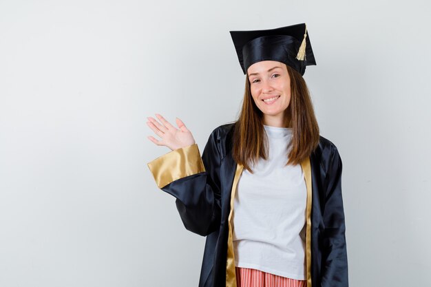 Laureato femminile agitando la mano per il saluto in uniforme, abbigliamento casual e guardando gioioso, vista frontale.