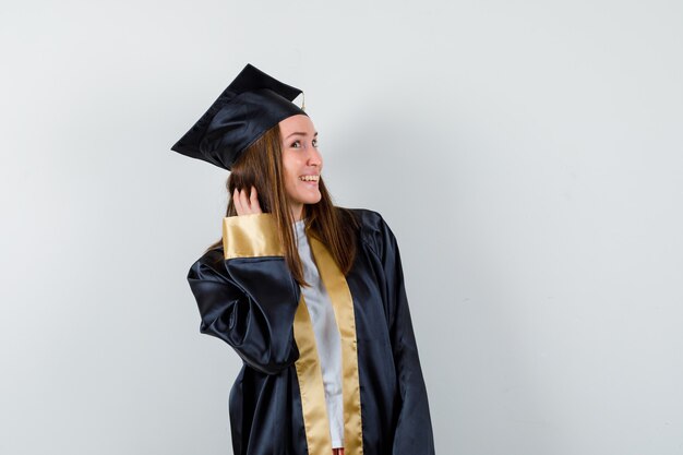 Laureato femminile giovane che tiene la mano sui capelli mentre posa in vestito accademico e sembra fresco. vista frontale.