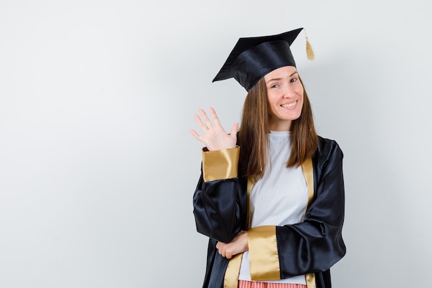 Laureato femminile in uniforme, vestiti casual agitando la mano per dire addio e guardando allegro, vista frontale.