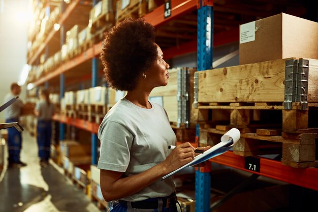 Lavoratore afroamericano che scrive l'elenco dell'inventario mentre controlla le scorte nel magazzino