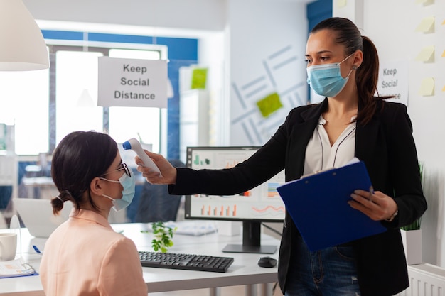 Lavoratore dell'azienda che mantiene la distanza sociale scansionando la temperatura del collega con il termometro a infrarossi per prevenire l'infezione da virus durante la pandemia globale con covid-19. Controllo sanitario dell'ufficio wo