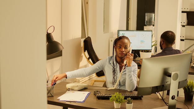 Lavoratore dell'azienda che risponde alla telefonata di rete fissa in ufficio durante il tramonto. Parlando su una linea di telecomunicazioni remota con il telefono cordless dell'ufficio, lavorando sulla presentazione della ricerca.