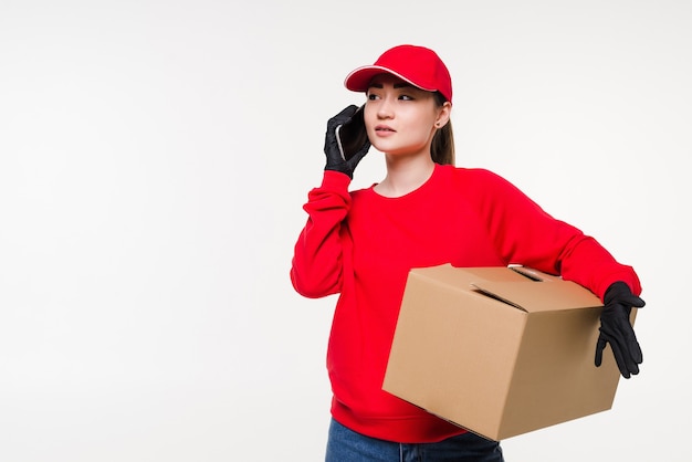 Lavoratore di consegna femminile giovane asiatico in piedi nel muro bianco utilizzando smartphone mobile parlando con il cliente che controlla l'indirizzo di spedizione